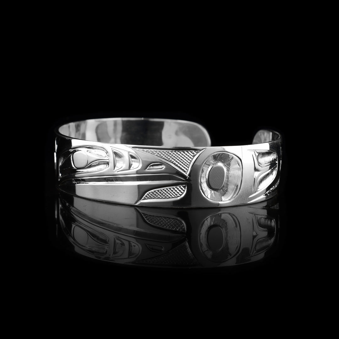 This Raven bangle bracelet has a single, sterling silver band with a gap in the back. The head of the Raven is shown in the front at a side profile. The head is large and stretches across most of the band. On either end of the band are wing designs and there are cross hatching designs in the background of the scene.