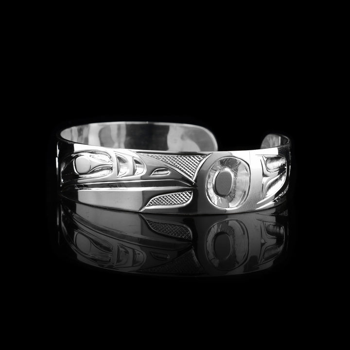 This Raven bangle bracelet has a single, sterling silver band with a gap in the back. The head of the Raven is shown in the front at a side profile. The head is large and stretches across most of the band. On either end of the band are wing designs and there are cross hatching designs in the background of the scene.