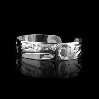This Raven bangle bracelet has a single, sterling silver band with a gap in the back. The head of the Raven is shown in the front at a side profile. The head is large and stretches across most of the band. On either end of the band are wing designs and there are cross hatching designs in the background of the scene.