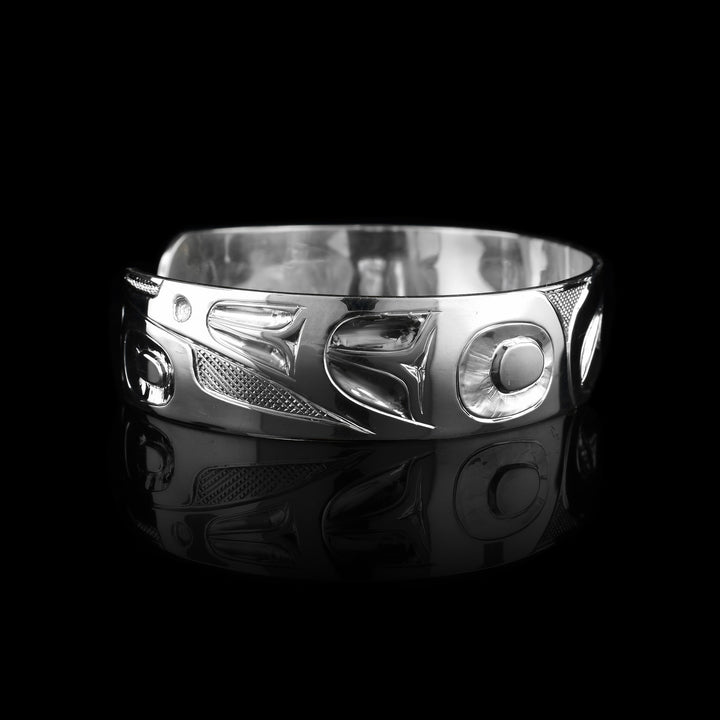 This Hummingbird bangle bracelet has a single, sterling silver band with a gap in the back. The head of the Hummingbird is shown in the front at a side profile. The head is large and stretches across most of the band. On either end of the band are wing designs and there are cross hatching designs in the background of the scene.