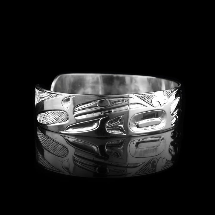 This Wolf bangle bracelet has a single, sterling silver band with a gap in the back. The head of the Wolf is shown in the front at a side profile. The head is large and stretches across most of the band. On either end of the band are paws and tail designs and there are cross hatching designs in the background of the scene.