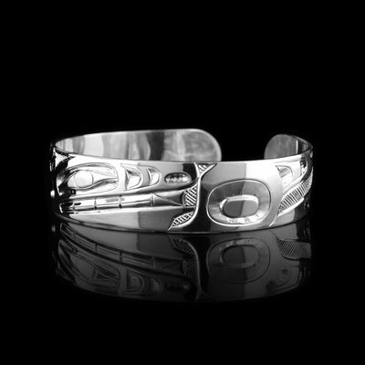 This Wolf bangle bracelet has a single, sterling silver band with a gap in the back. The head of the Wolf is shown in the front at a side profile. The head is large and stretches across most of the band. On either end of the band are paws and tail designs and there are cross hatching designs in the background of the scene.