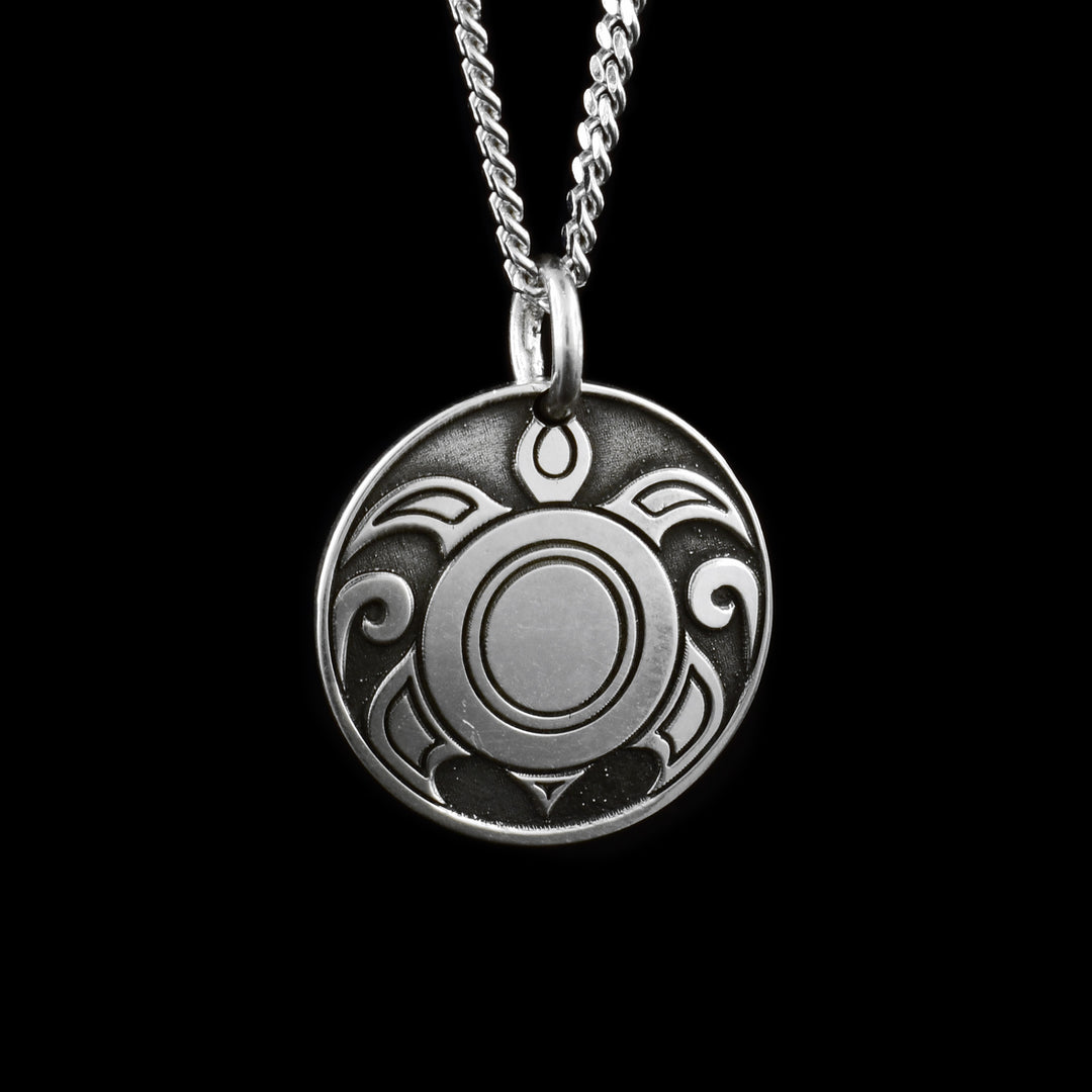 This sea turtle pendant necklace has a small, circle shape and is made from sterling silver. There is a tribal, like depiction of a turtle from above with swirling water designs in the background.