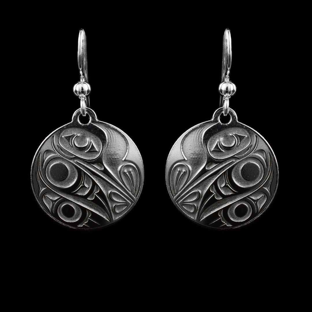 These small dangle earrings have circle shaped hangs made form sterling silver. There is a depiction of the Hummingbird cast into the hangs at a side profile. The beak is facing down and there is one, large eye on the head. its wing and tail are tucked underneath is and its beak is sticking into a flower.