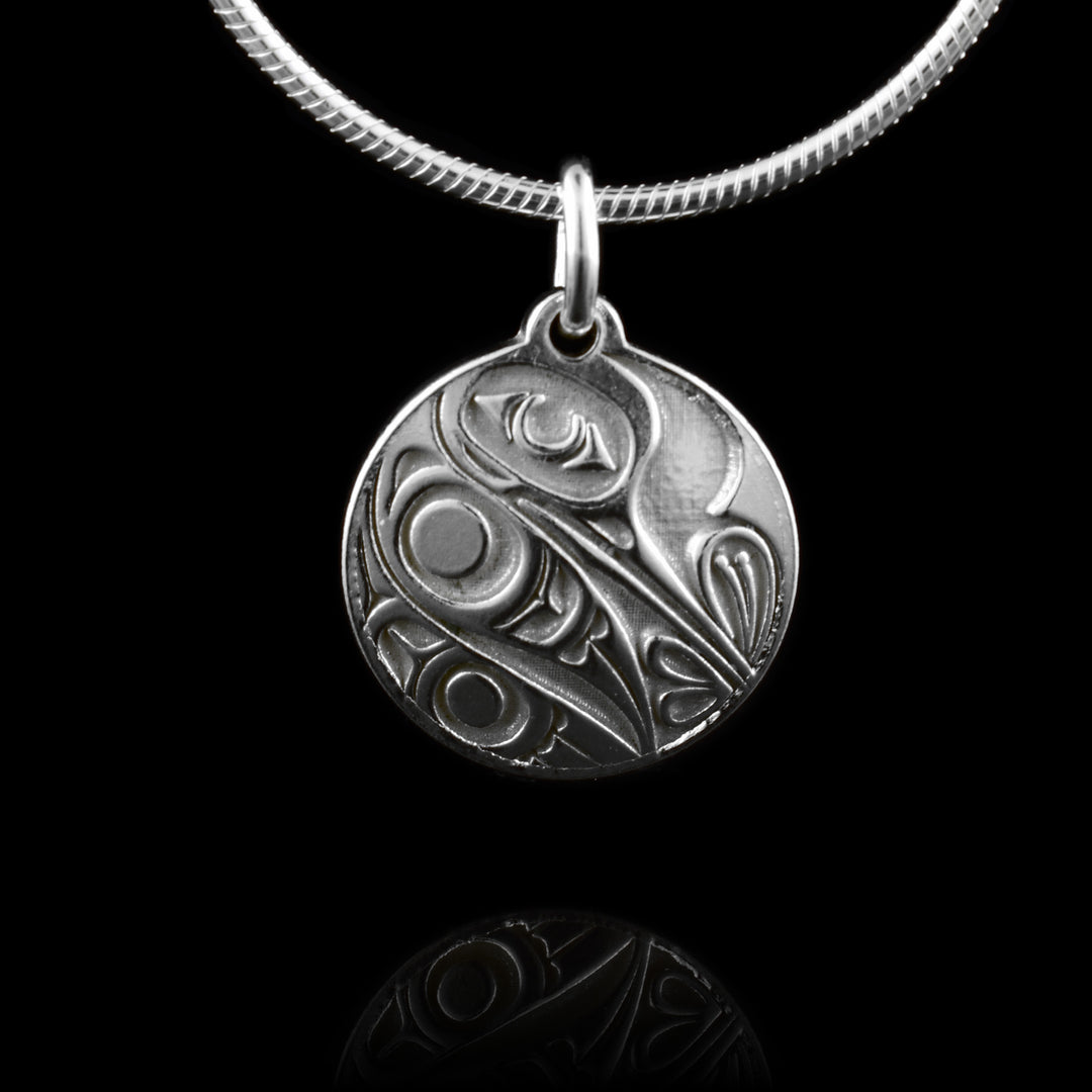 This Hummingbird silver pendant has a circle shape made from sterling silver. There is a depiction of the Hummingbird cast into the pendant at a side profile. The beak is facing down and there is one, large eye on the head. its wing and tail are tucked underneath is and its beak is sticking into a flower. 