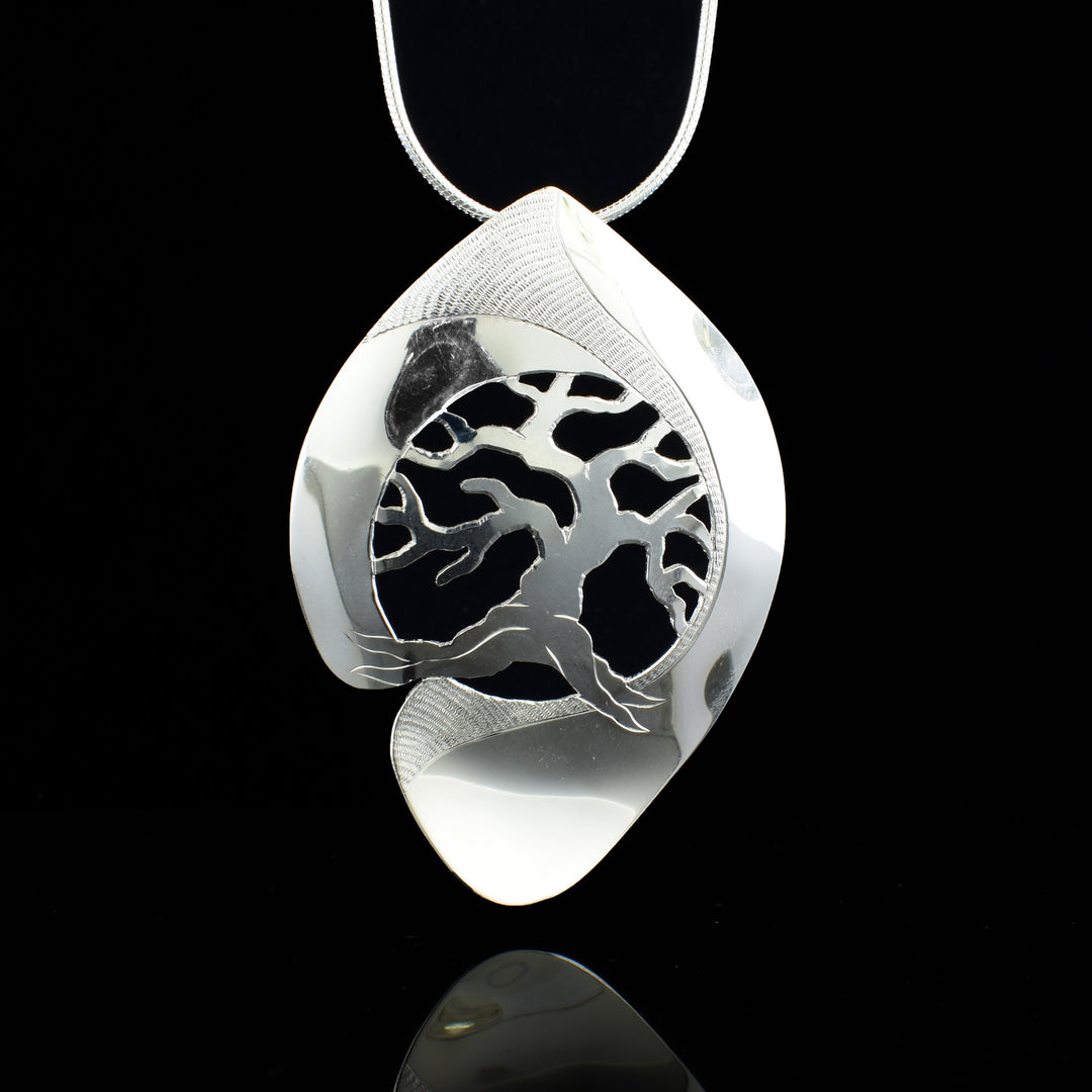 This tree of life pendant is large and made from sterling silver. The piece has a long, oblong shape with a circle cutout in the center. There are cutout designs detailing a sprawling tree in the center cutout. There are carvings on the piece detailing the scene.
