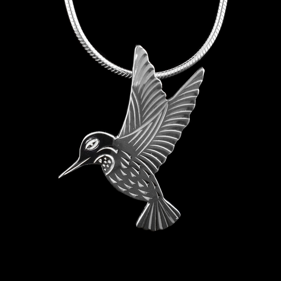 This hummingbird silver pendant is made form sterling silver and has been cutout to depict a Hummingbird in flight. Its beak is facing downward and its two wings are outstretched above it. There are eye and beak details carved as well as designs on the throat. There are feather designs across its body, wings and tail.
