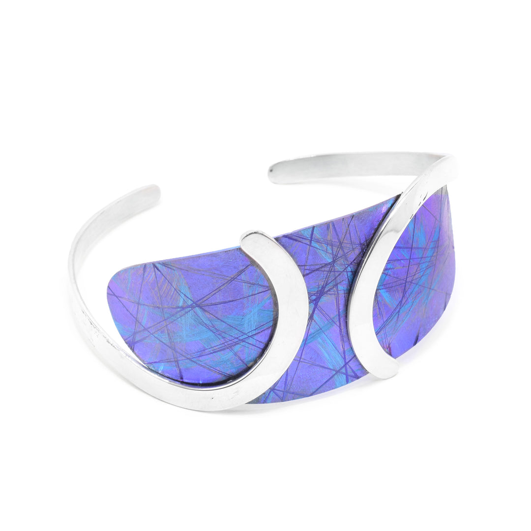 This blue titanium bracelet has a wide, oval piece at the top with a blue color and purple, line designs on it. This piece is held with swirling bands made from sterling silver. There is a gap between the silver bands at the bottom.