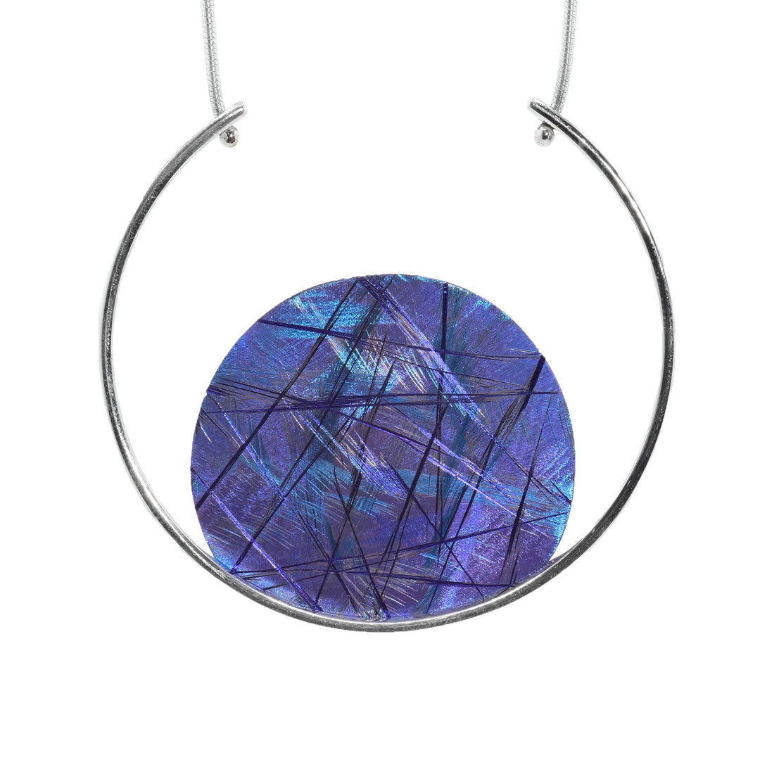 This titanium pendant necklace a sterling silver wire shaped like a circle. There is a circle pendant made from titanium that is resting in the wire. There are blue and purple designs across the circle titanium piece. The chain is connected on both sides of the circle wire.