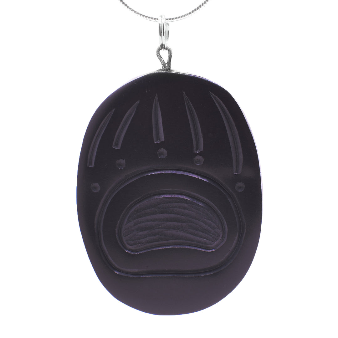 This Bear paw pendant has a large oval shape and is made from argillite. There are deep carvings made on its surface depicting a bears paw. The palm is large and the claws are long as the toes are small circles.