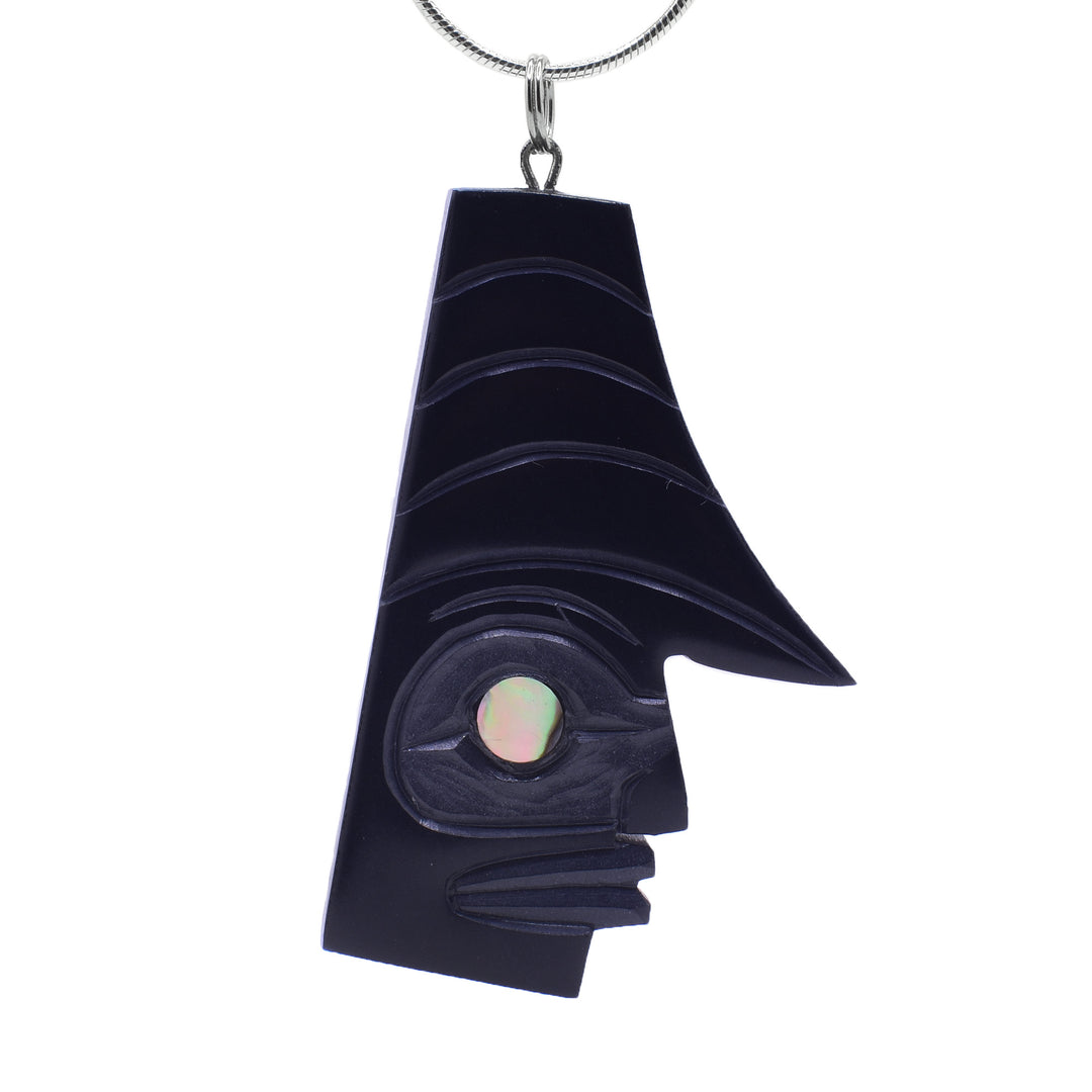 This argillite pendant has a long, rectangle shaped and the face of the "watchman" is carved into the bottom of the piece at a side profile. On one side, there is a large swoop that goes out above the eye. There is a circle abalone piece set in the eye.