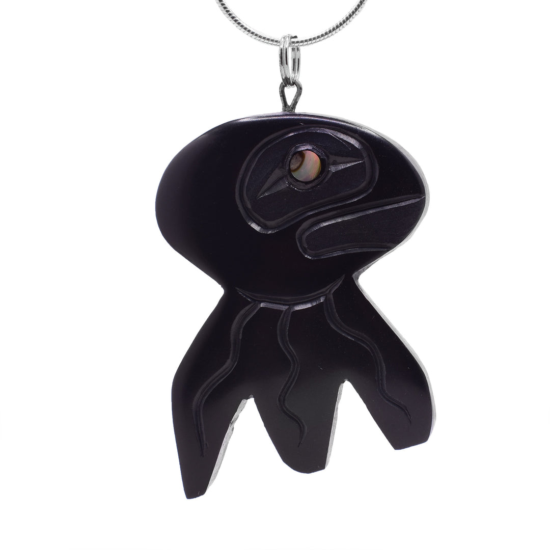 This unique jellyfish pendant is large and has been carved from argillite stone. The head is a wide, oval shape and there are three tentacles carved into the piece There are carvings on the surface of the pendant detailing the head, face and tentacles of the jellyfish.