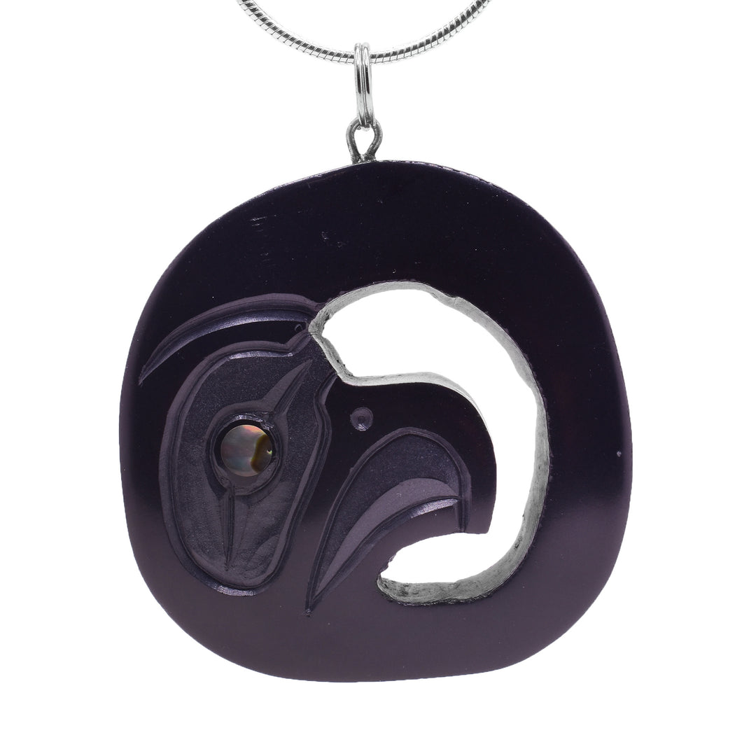 This Haida Eagle pendant is circle shaped and has been carved from argillite stone. There is a cutout around the head of the Eagle at a side profile in the center of the piece. There are carvings on its surface detailing the eye, nose and beak of the Eagle. In its eye there is an abalone gem set.