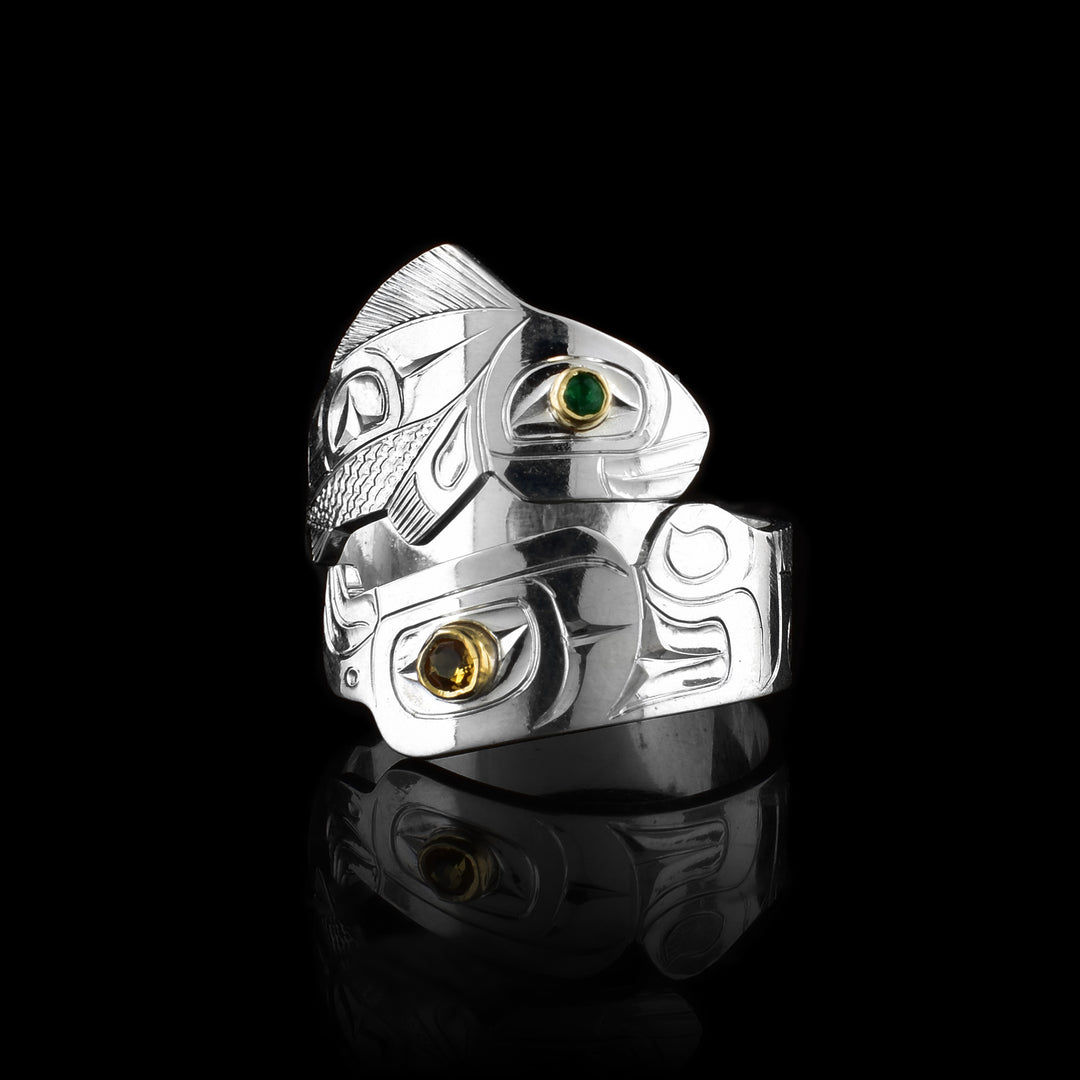 This multi gemstone ring has a sterling silver band that wraps around the finger twice. One band is cutout and carved to depict the salmon and the other depicts the eagle. There is an emerald set in gold in the salmon's eye and a citrine gem for the eagle.