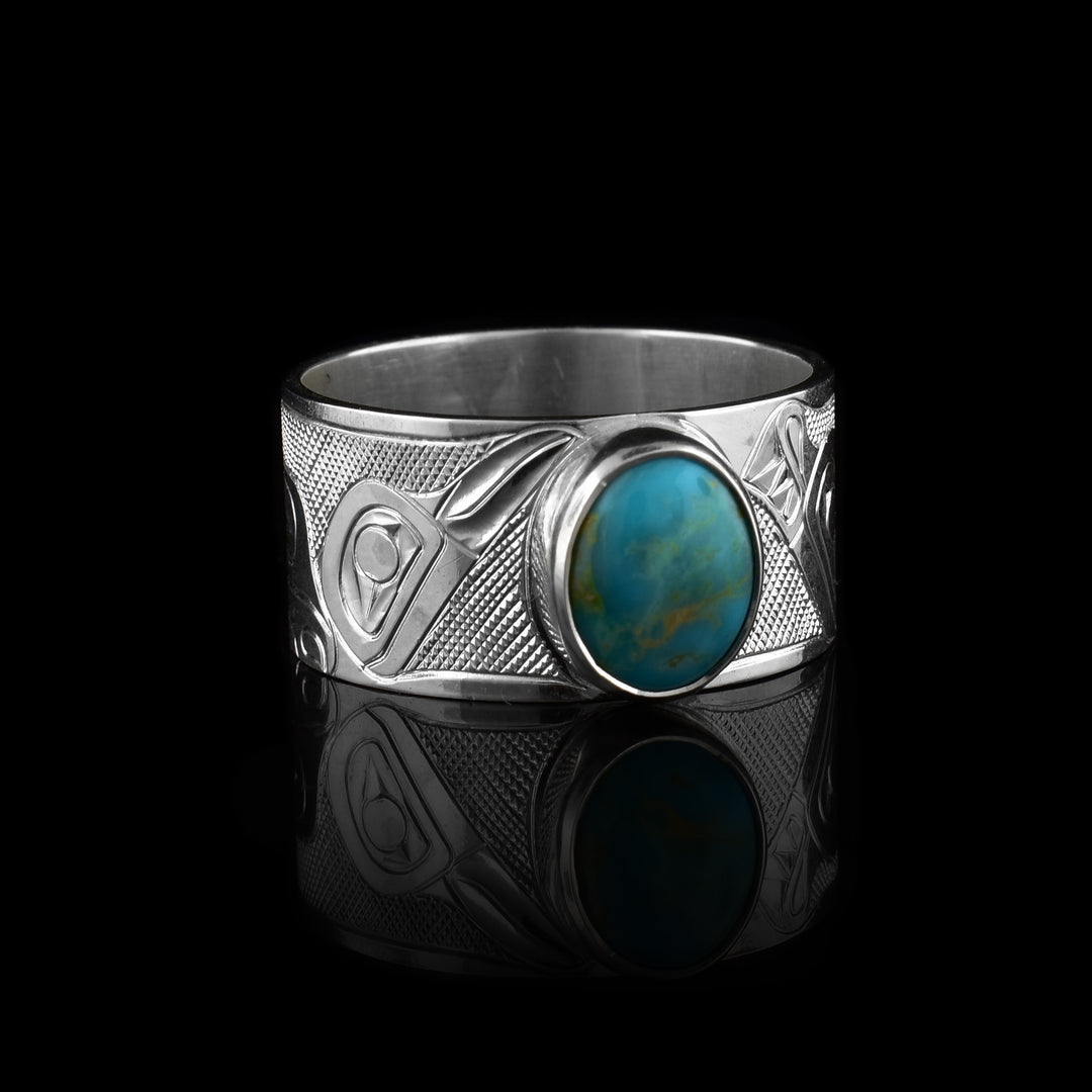 This wide band solitaire ring has a large band made from sterling silver and there are side profile depictions of the heads of the Wolf and raven legend carved into it. There is a large, oval turquoise stone set in the silver. 