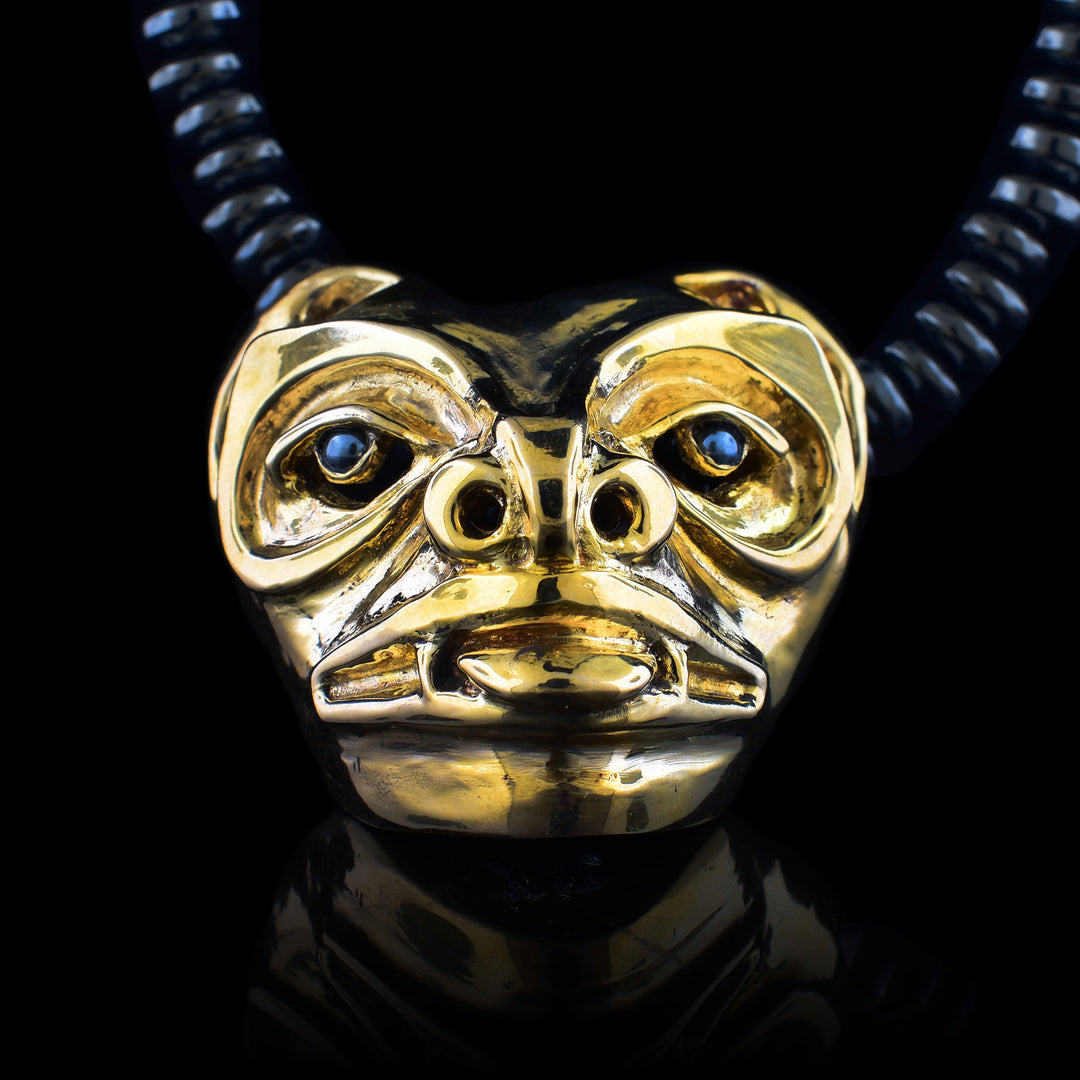 This bronze pendant depicts a Bear and is shaped like a mask with 3D details. Its eyes, eyebrows, nose, mouth and tongue are detailed. There are hematite stone held in the eyes. It is on a beaded necklace with round, flat hematite beads.