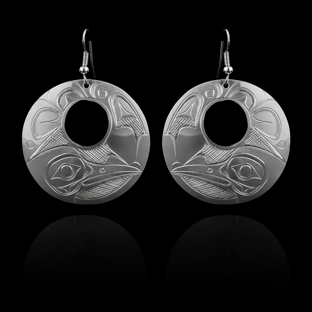 These large silver earrings have sterling silver  hangs with a circle shape and small, circle cutout in the center of them. The head of the Raven is depicted at the bottom in a side profile. The legend has a long beak and there are wide, feather designs carved around the rest of the piece.