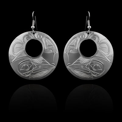 These large silver earrings have sterling silver  hangs with a circle shape and small, circle cutout in the center of them. The head of the Raven is depicted at the bottom in a side profile. The legend has a long beak and there are wide, feather designs carved around the rest of the piece.