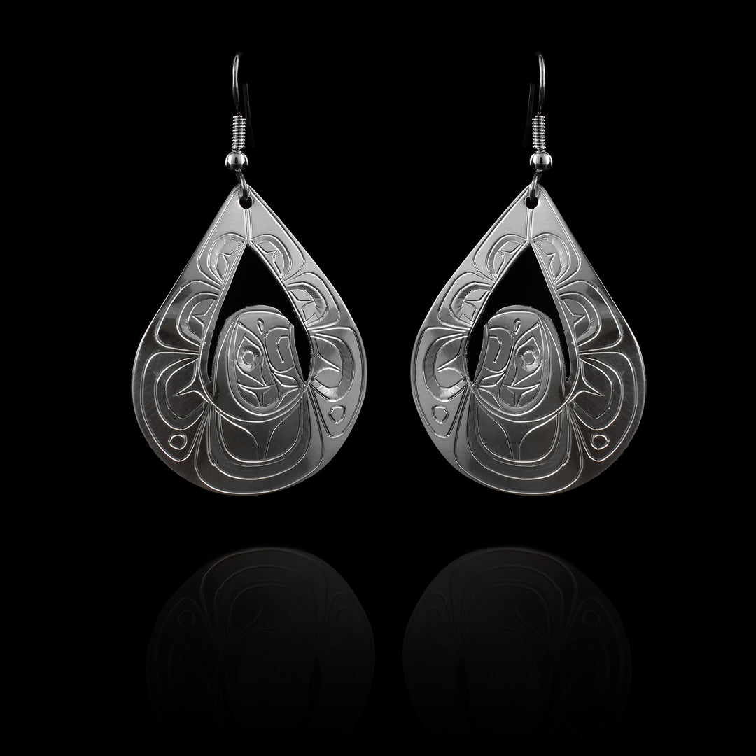 These large silver earrings have wide, teardrop shaped hangs with cutouts in the center of them. In the center of the hangs is The head of the Eagle and its wings outstretched above its head to form the teardrop shape. There are carvings on the surface of the hangs depicting the Eagle.