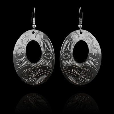 These large silver earrings have wide, oval shaped hangs with small, oval cutouts in the center of them. The head of the Raven is carved into the bottom with a side profile. There are feather designs made around the rest of the piece.