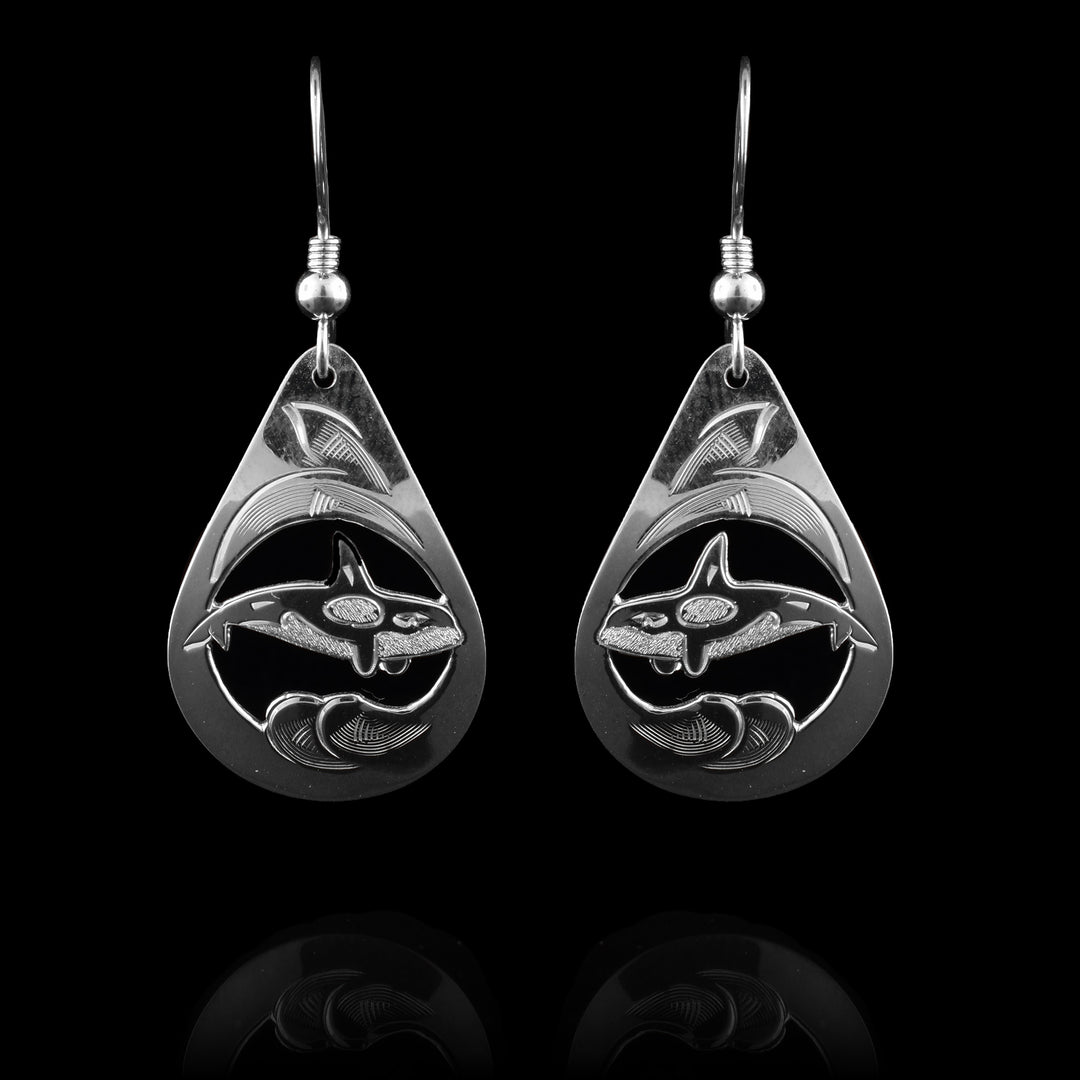 These dangle Orca whale earrings have teardrop shaped hangs that are made from sterling silver. There are cutout depictions of the Orca whale in the center of the hangs with wavy, ocean designs around them. The area around the whales is cutout and there are hatching designs detailing the orcas.