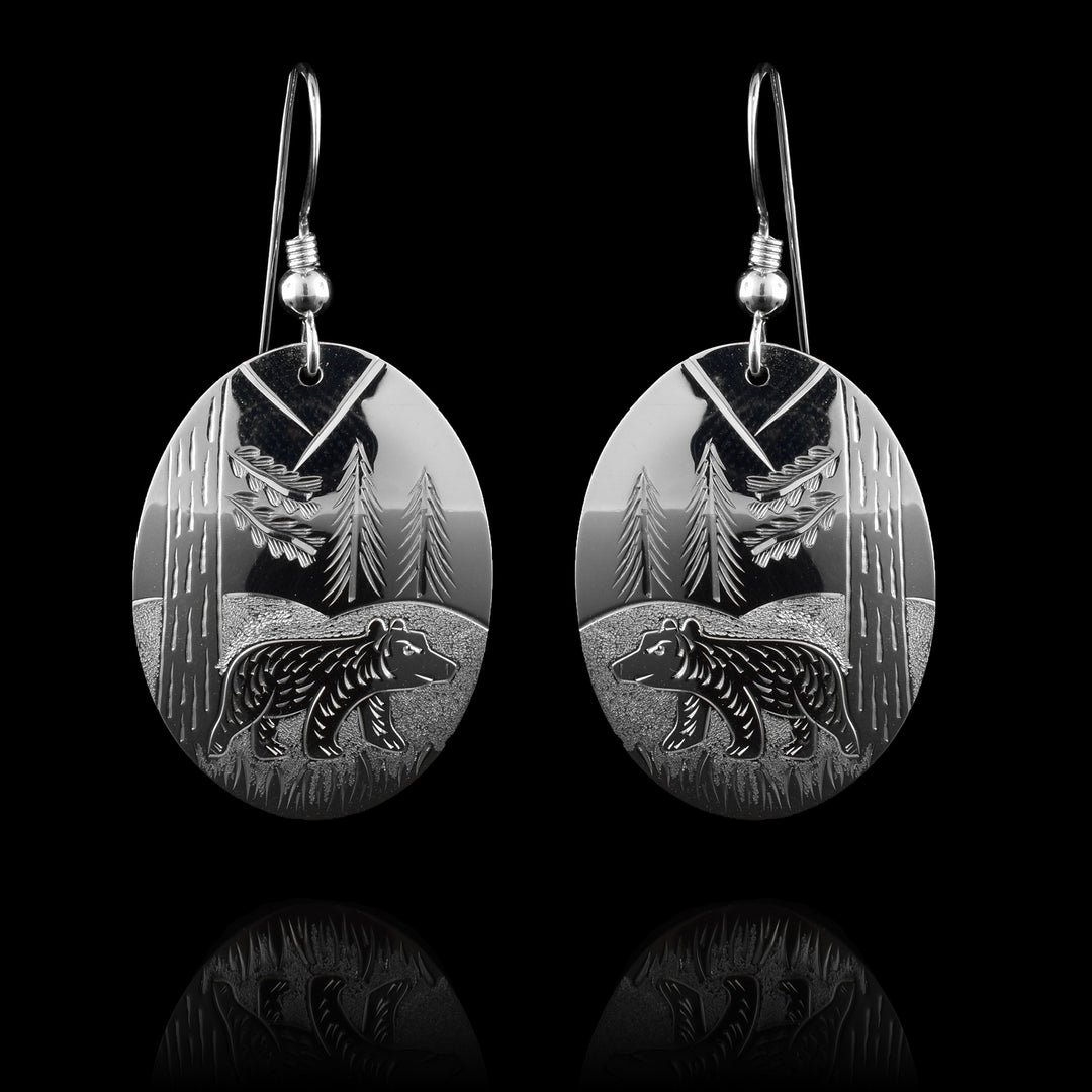 These bear nature earrings have oval shaped hangs and are made from sterling silver. There are carings on the hangs showing a bear at a side profile with hills and trees detailed around it.