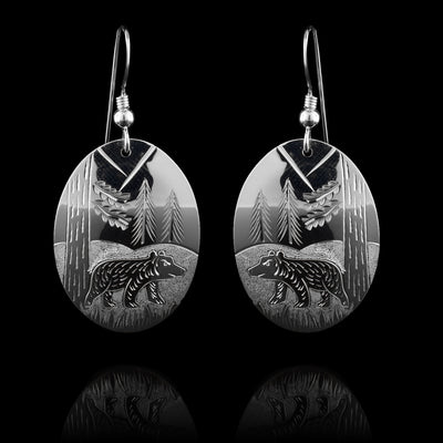 These bear nature earrings have oval shaped hangs and are made from sterling silver. There are carings on the hangs showing a bear at a side profile with hills and trees detailed around it.