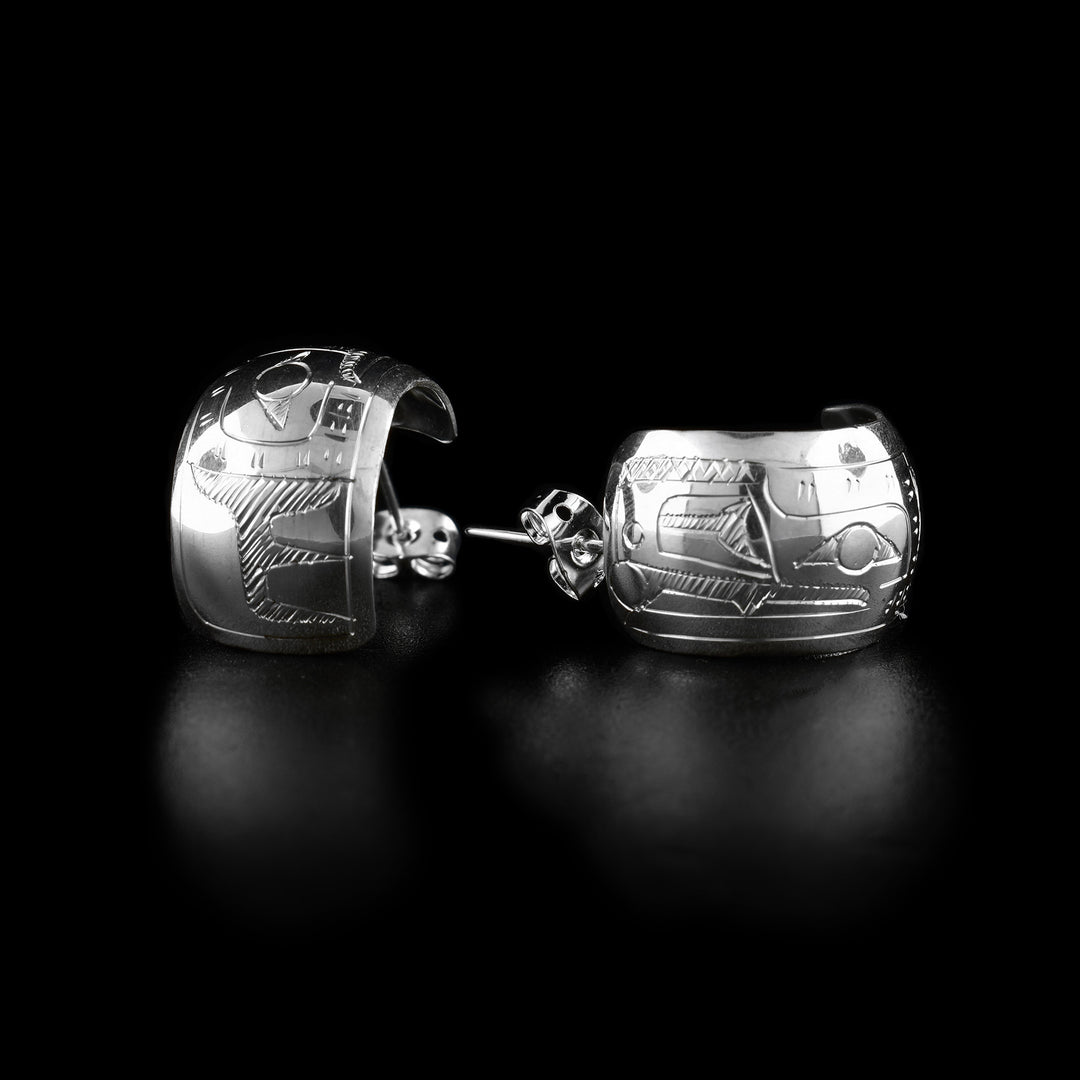 These wide hoop earrings are made from sterling silver and have a single, wide band with a gap in the back. There are carvings on the surface of the hoops depicting the Raven. The head of the Raven is facing the studs and there is a pearl depicted in the mouth of the legend.