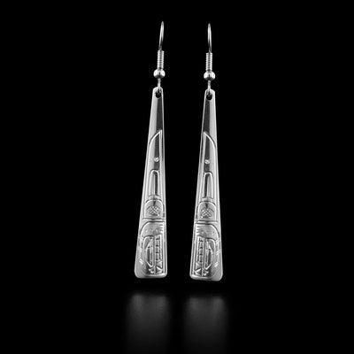 These drop triangle earrings are made from sterling silver and have long, thin triangle shaped hangs. There are depictions of the Orca legend carved into the surface of the hangs depicted at a side profile. The head is at the bottom and there is a dorsal fin design behind the head.