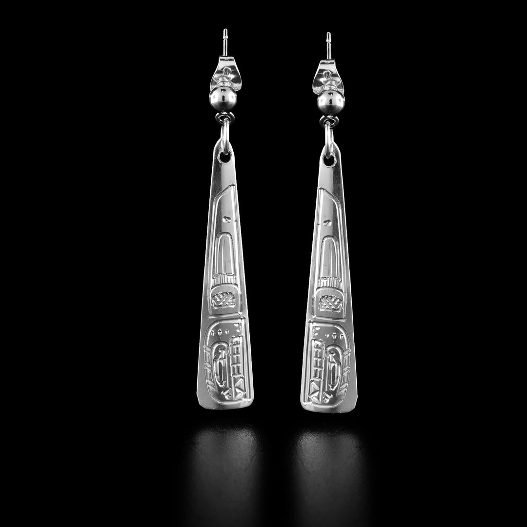 These drop triangle earrings are made from sterling silver and have long, thin triangle shaped hangs. There are depictions of the Orca legend carved into the surface of the hangs depicted at a side profile. The head is at the bottom and there is a dorsal fin design behind the head.