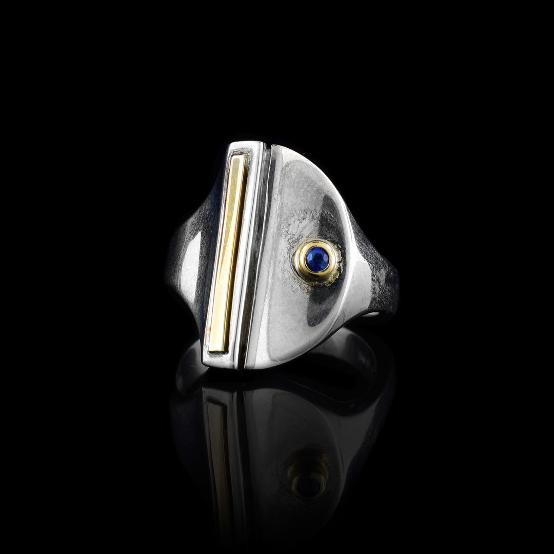this sapphire signet ring has a large, oval signet made from sterling silver. There is a 14K gold bar running through the center of the signet. On one side of the signet, there is a small sapphire gem set with 14K gold.
