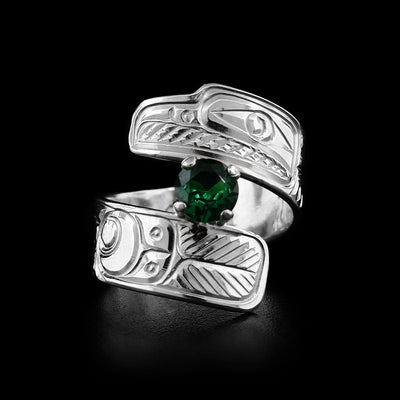This silver wrap ring has a dark green lab grown emerald in a silver setting in the center. The top part of the wrap ring depicts the head of a raven facing the left and the remainder of the band has intricate carvings representing feathers.