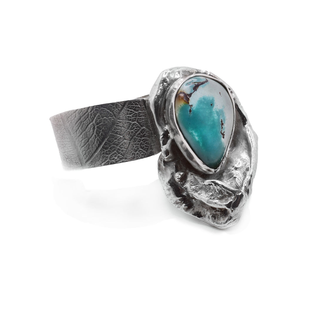 This turquoise ring has a wide, silver band and a unique, leaf-like shape of silver encasing the teardrop shaped turquoise slightly off-center. When worn, the turquoise covers the adjacent finger.