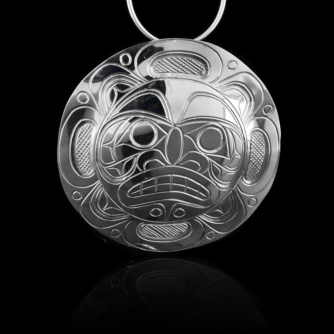 This large Moon pendant is made from sterling silver and has a large, circle shape. The face of the Moon legend is carved into the center of the piece with its eyes, nose and teeth detailed. around the face, there are design depicting the moon.