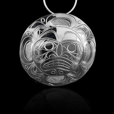 This large Moon pendant is made from sterling silver and has a large, circle shape. The face of the Moon legend is carved into the center of the piece with its eyes, nose and teeth detailed. around the face, there are design depicting the moon.