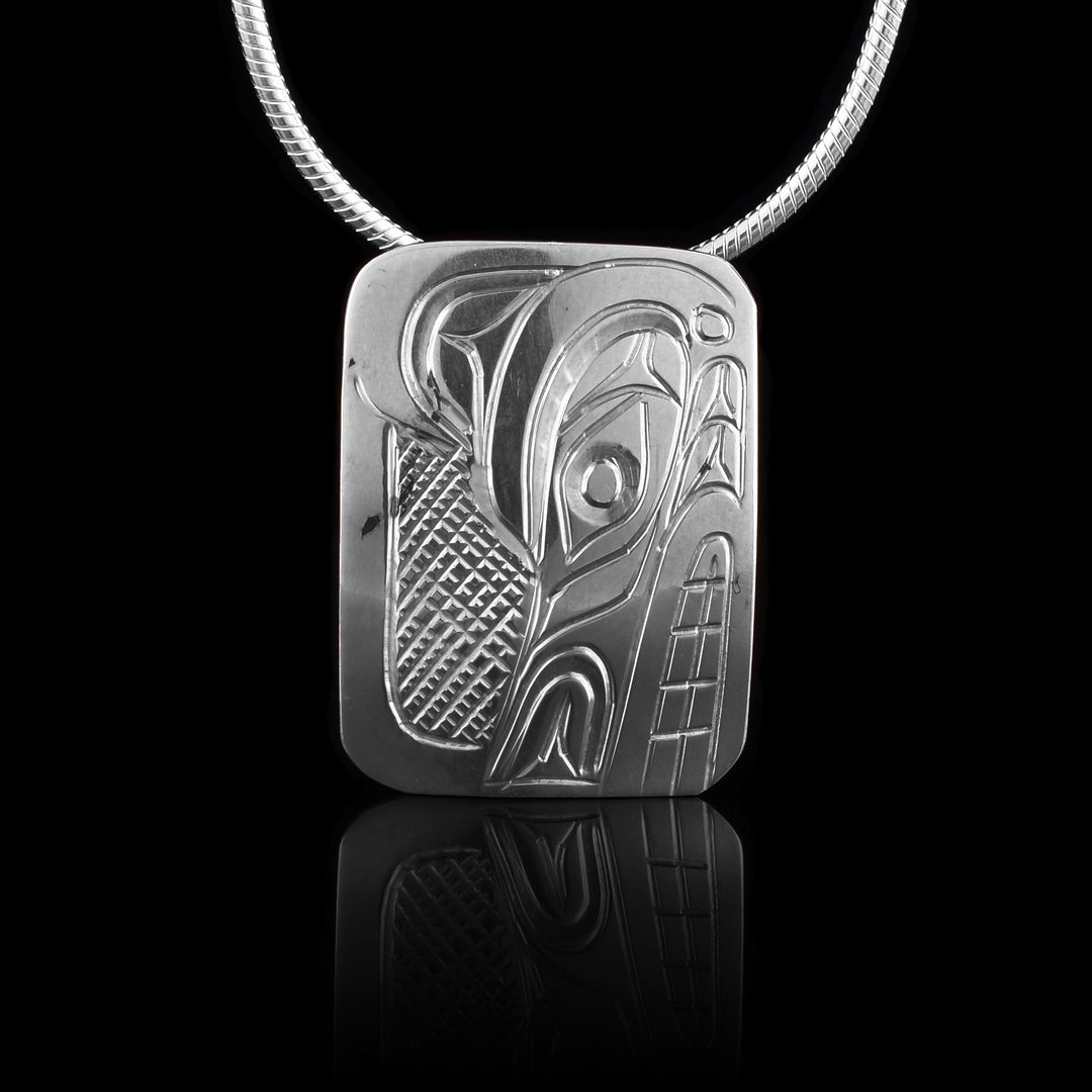 This Wolf head pendant is made from sterling silver and has a small rectangle shape. There is a depiction of the head of the Wolf carved in a side profile on the piece. Its snout is facing down and there are designs on its face. There are cross hatching designs in the background of the scene.