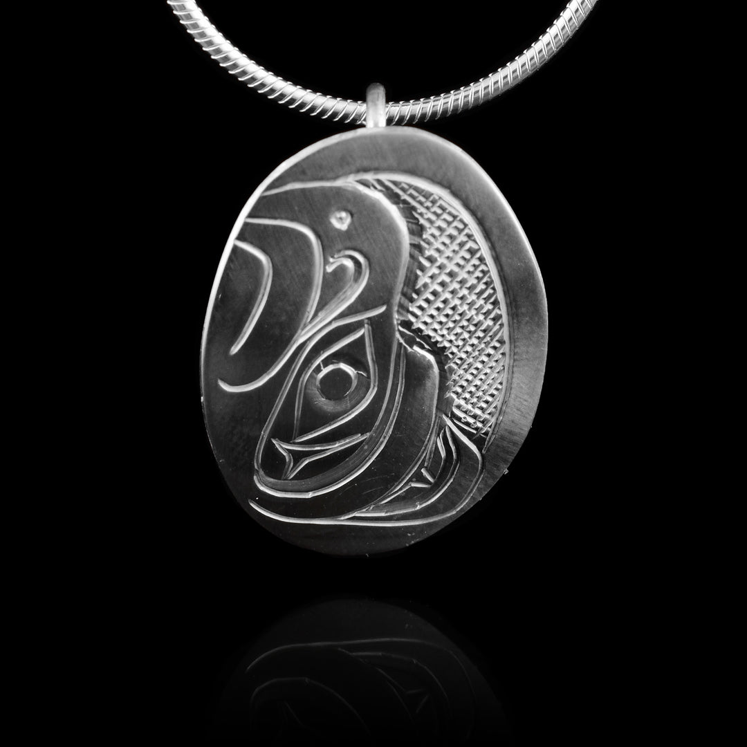 This Thunderbird head pendant is made from sterling silver and has a small oval shape. There is a depiction of the head of the Thunderbird carved in a side profile on the piece. Its beak is facing up and there are designs on its face. There are cross hatching designs in the background of the scene.