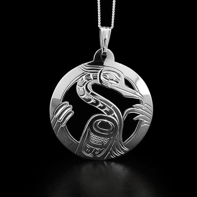 This heron pendant is round in shape and depicts a full-bodied heron facing the left when worn. There are cattails and leaves surrounding it. The background has been cut out for depth.