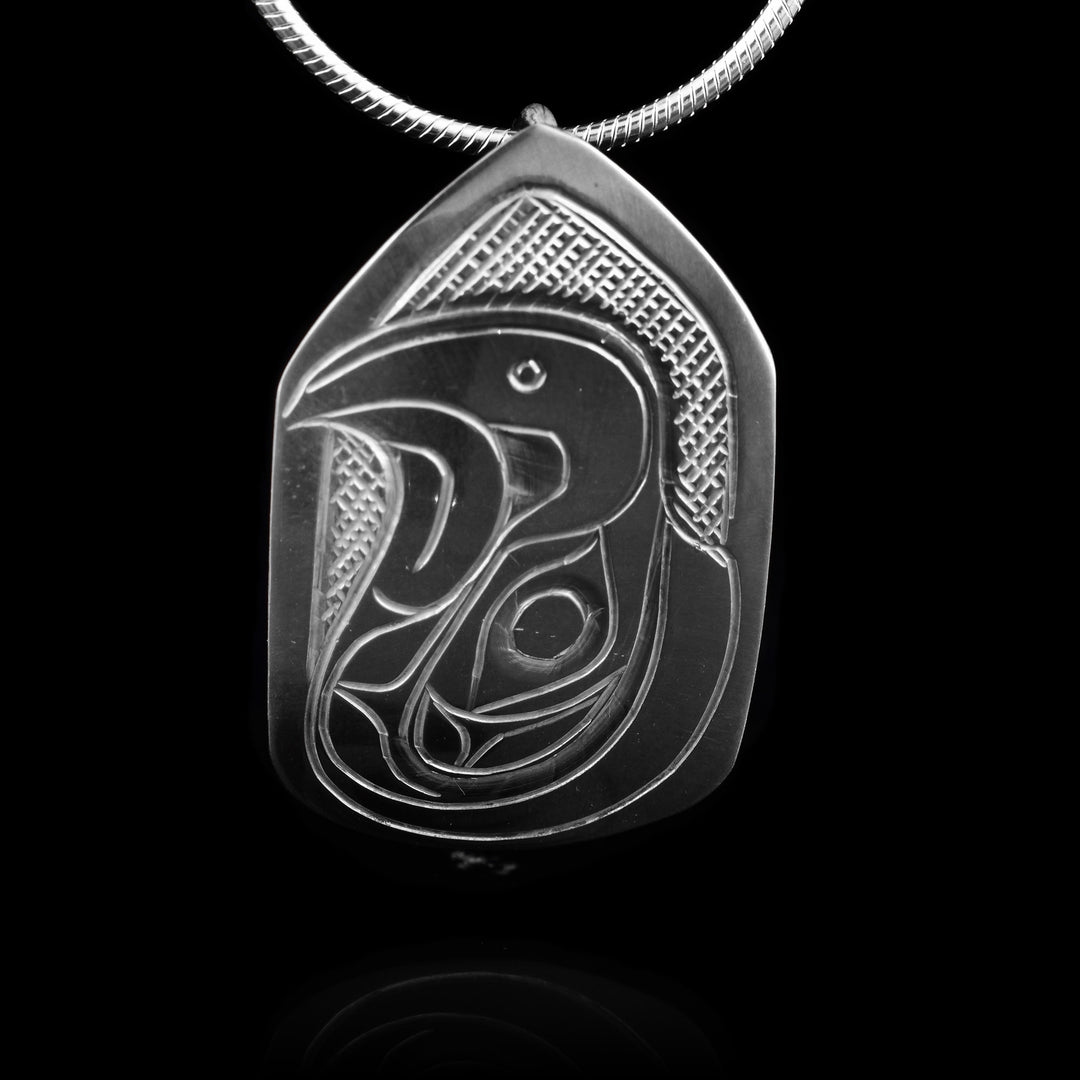 This Eagle head pendant is made from sterling silver and has a small pentagon shape. There is a depiction of the head of the Eagle carved in a side profile on the piece. Its curved beak is facing up and there are designs on its face. There are cross hatching designs in the background of the scene.