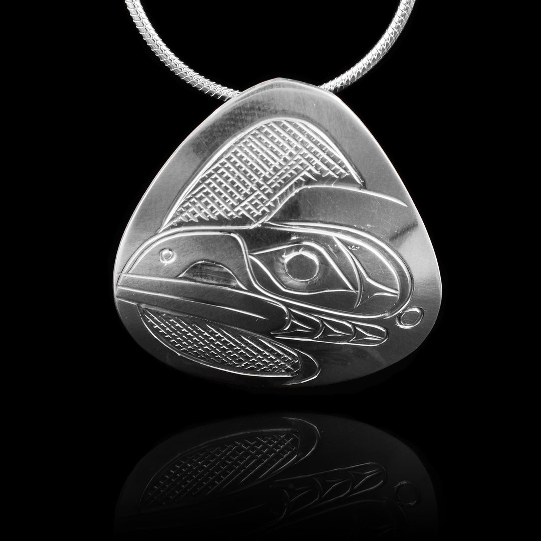 This Raven head pendant is made from sterling silver and has a small, wide teardrop shape. There is a depiction of the head of the Raven carved in a side profile on the piece. Its beak is facing towards the side and there are designs on its face. There are cross hatching designs in the background of the scene.
