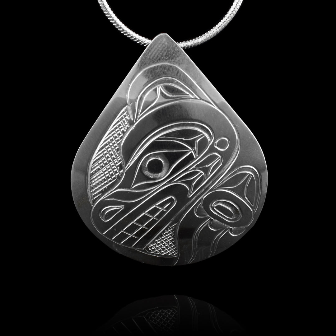 This Bear head pendant is made from sterling silver and has a small, wide teardrop shape. There is a depiction of the head of the Bear carved in a side profile on the piece. Its snout is facing down towards the side and there are designs on its face. There are cross hatching designs in the background of the scene.