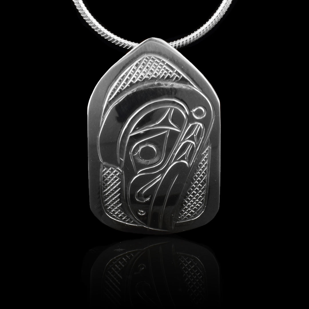 This Raven head pendant is made from sterling silver and has a small pentagon shape. There is a depiction of the head of the Raven carved in a side profile on the piece. Its beak is facing down and there are designs on its face. There are cross hatching designs in the background of the scene.