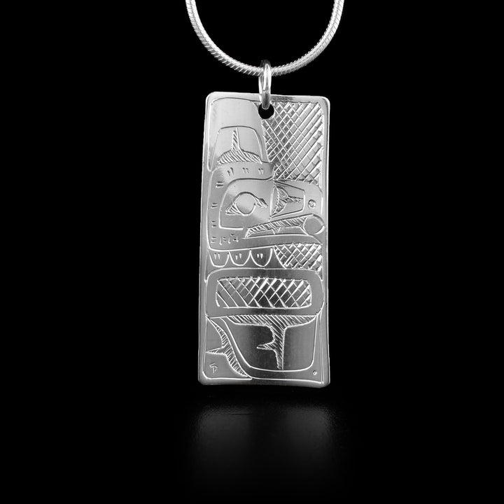 This Raven and Bear pendant is double sided and this side shows the Raven. The Raven is depicted in a side profile with a large feather detail on the top of its head and a pearl is shown being held in its beak. Its wing is shown at the bottom of the piece and there are cross hatching designs in the background of this scene.