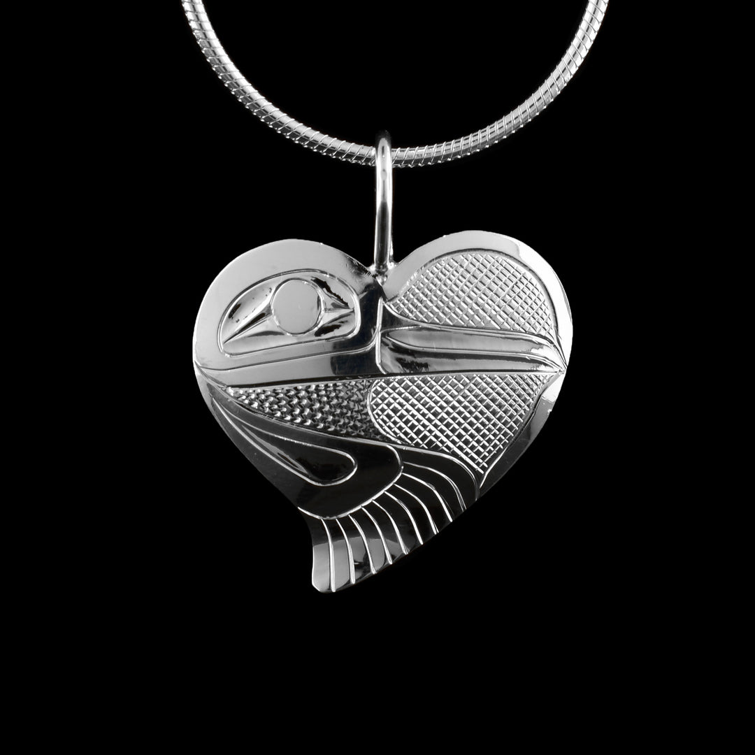 This sterling silver heart pendant necklace  is heart shaped and has a depiction of the Hummingbird carved into it. The bird is in one side of the piece and has its beak going across the piece. There are cross hatching designs made in the background of the scene. There is a wing curled underneath the head with feather designs.