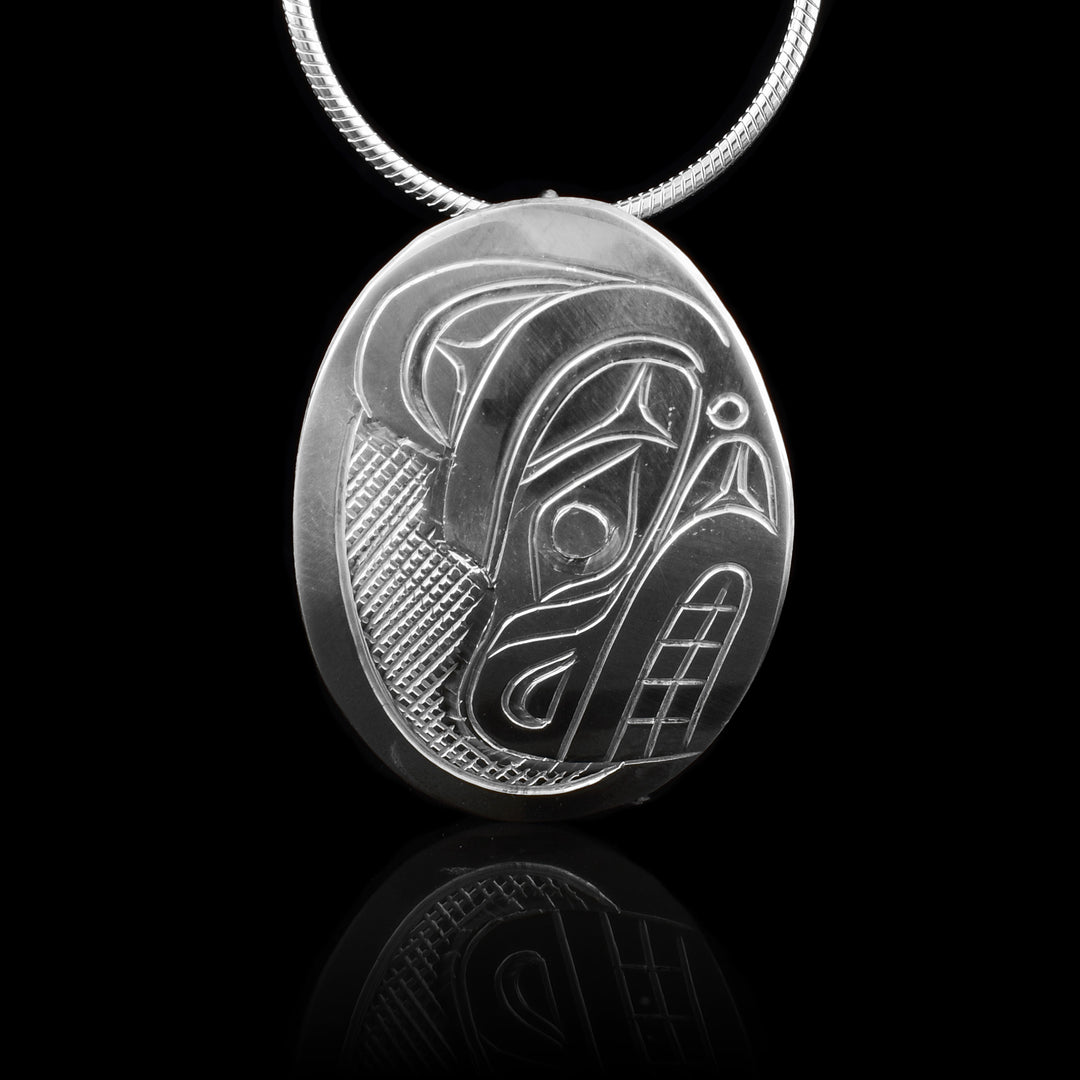 This Bear head pendant is made from sterling silver and has a small oval shape. There is a depiction of the head of the Bear carved in a side profile on the piece. Its snout is facing down and there are designs on its face. There are cross hatching designs in the background of the scene.