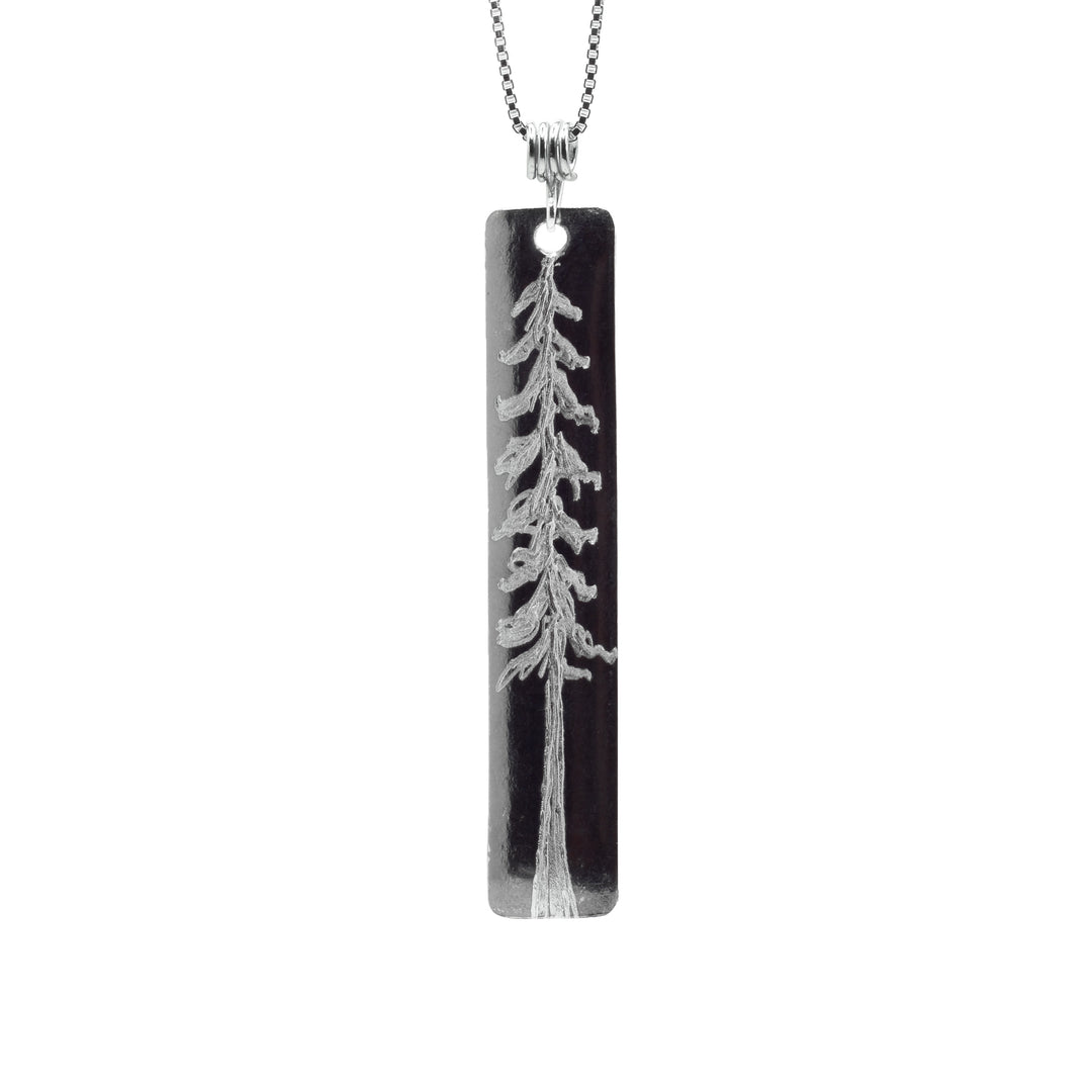 This cedar tree pendant is made from sterling silver and has a long, thin rectangle shape. There is a depiction of a tall, cedar tree carved into the surface of the piece.