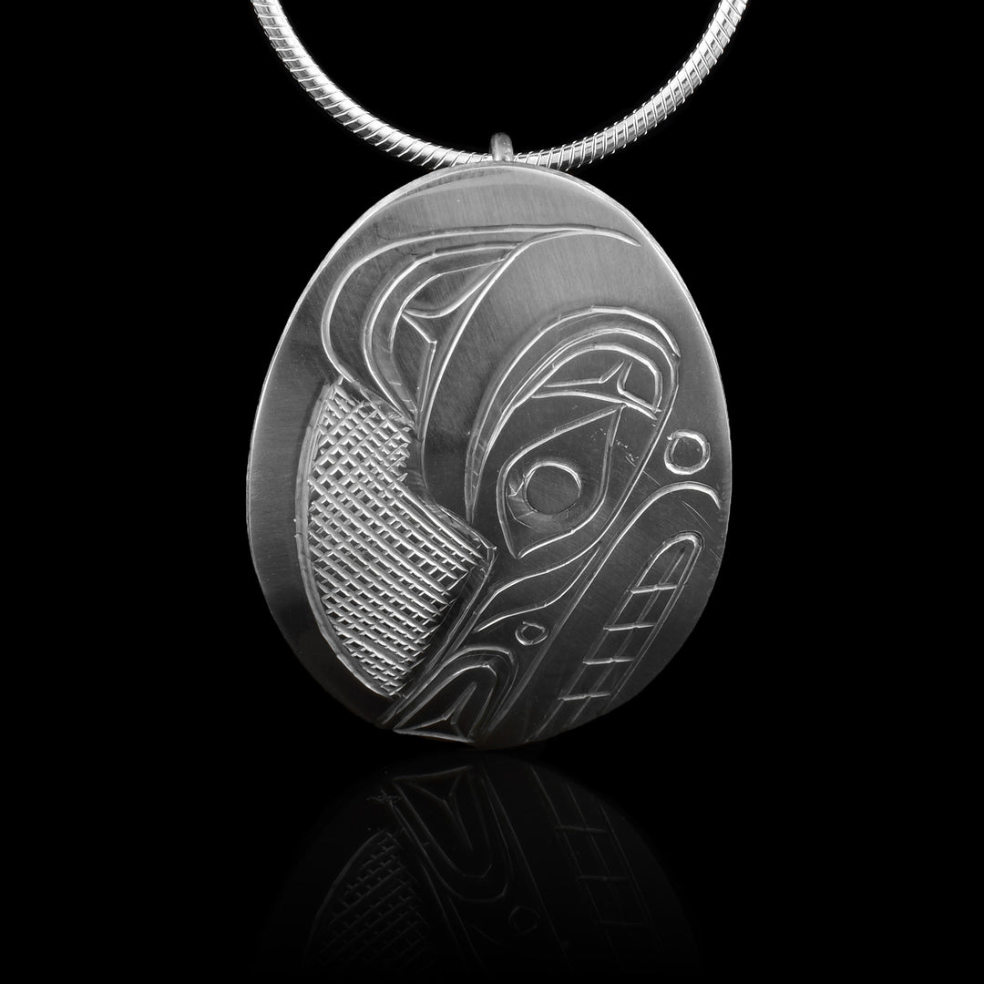 This Wolf head pendant is made from sterling silver and has a small oval shape. There is a depiction of the head of the Wolf carved in a side profile on the piece. Its snout is facing down and there are designs on its face. There are cross hatching designs in the background of the scene.