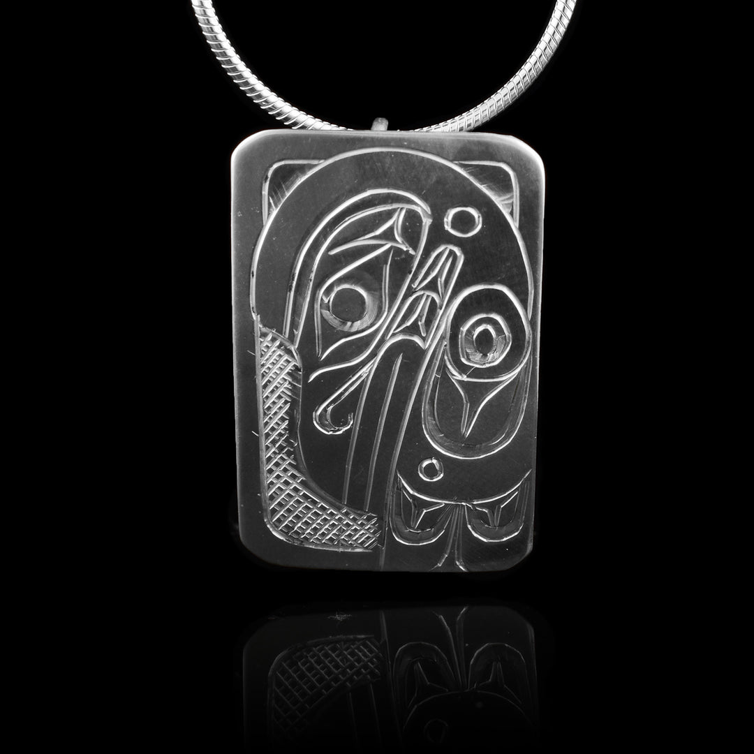 This Raven head pendant is made from sterling silver and has a small rectangle shape. There is a depiction of the head and wing of the Raven carved in a side profile on the piece. Its beak is facing down and its wing runs along the back of the beak. There are cross hatching designs in the background of the scene.