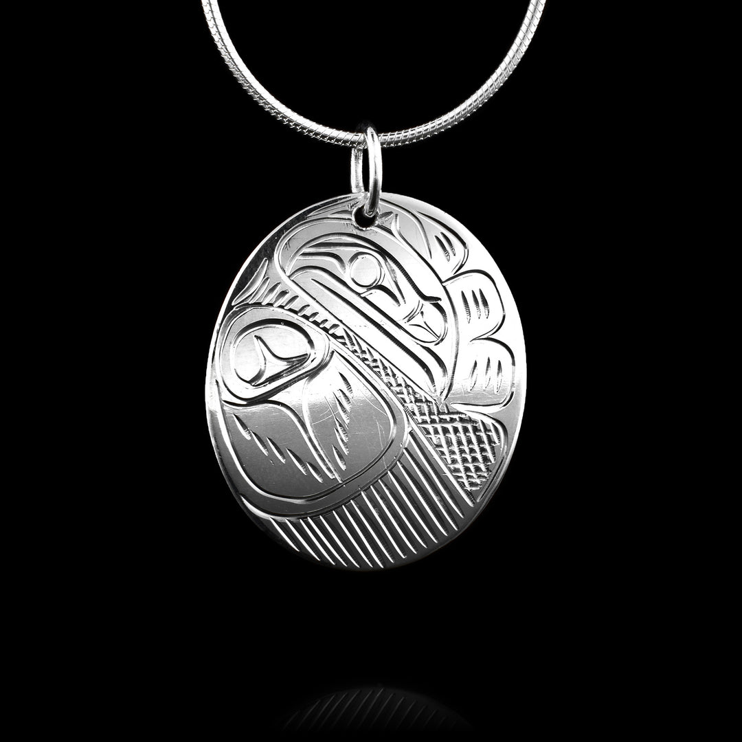 This silver pendant is oval in shape and depicts the Raven. The head of the legend with its feathers and curved beak is at the top with a large, spread out wing underneath. Cross-hatching has been carved to depict the negative space.