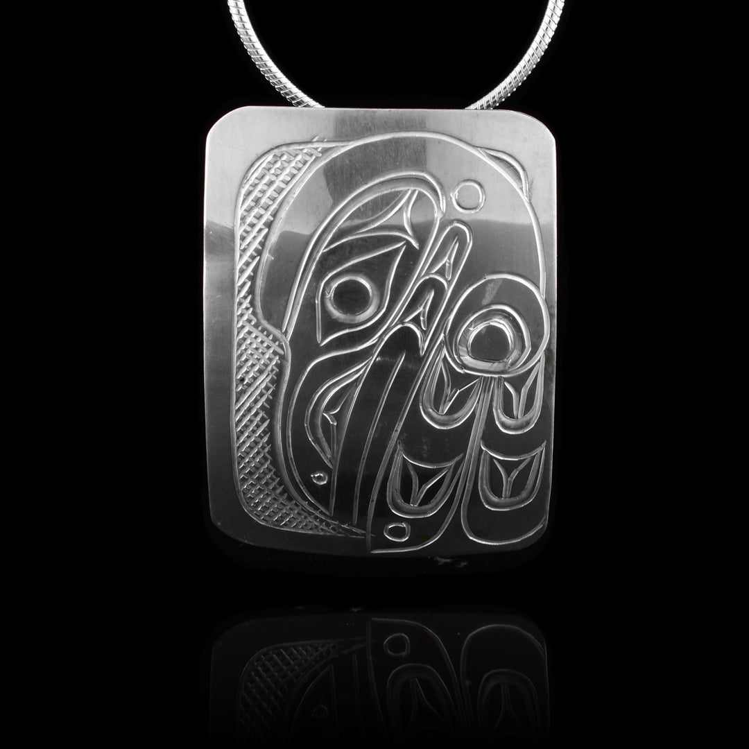 This Raven head pendant is made from sterling silver and has a wide rectangle shape. There is a depiction of the head and wing of the Raven carved in a side profile on the piece. Its beak is facing down and its wing runs along the back of the beak. There are cross hatching designs in the background of the scene.